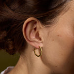 A close-up of a person's ear wearing a small textured gold hoop earring, with brown hair pulled back and a soft olive background.