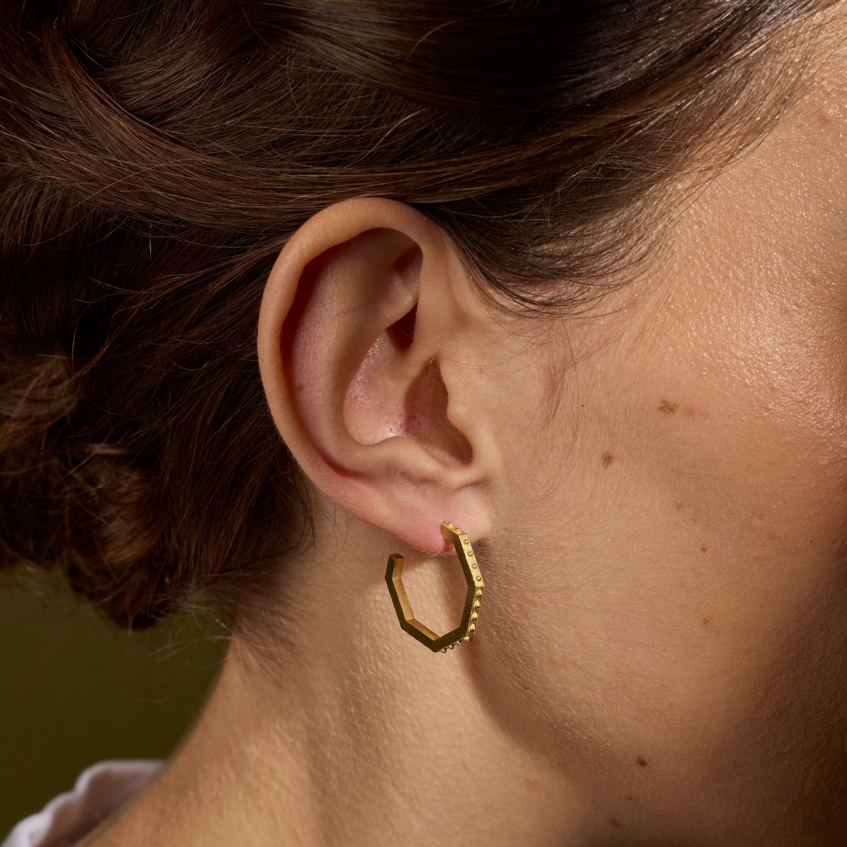 A close-up of a person's ear wearing a small textured gold hoop earring, with brown hair pulled back and a soft olive background.